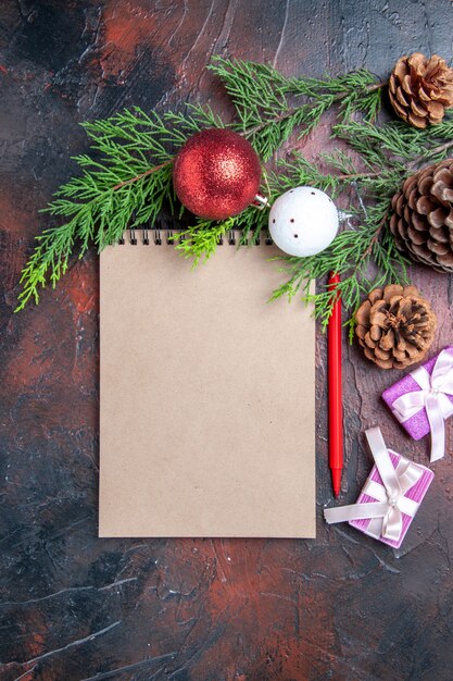 Vista superior lápiz rojo un cuaderno ramas de árboles de pino juguetes y regalos de bolas de árbol de navidad en la superficie de color rojo oscuro