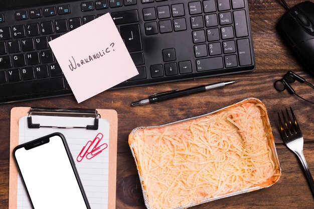 Vista superior lasaña y teclado con cuaderno en blanco y teléfono