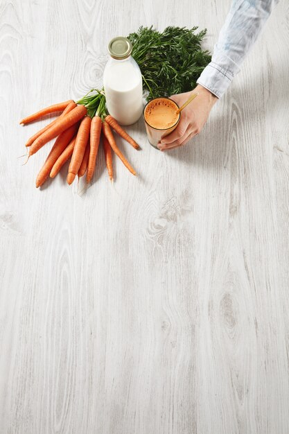 Vista superior lateral de la mesa de madera, cosecha de zanahoria de la granja que se encuentra cerca de la botella y la mano del hombre sostiene un vaso lleno de una mezcla de jugo fresco natural y leche con una pajita dorada para beber