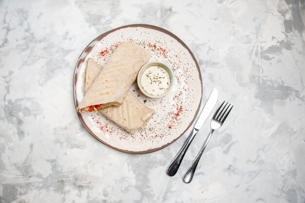 Vista superior de lavash wrap y yogur en un tazón pequeño sobre un plato y cubiertos sobre una superficie blanca manchada