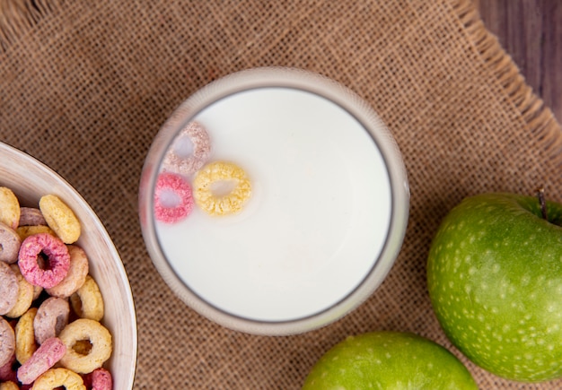 Vista superior de la leche en un tazón con cereales coloridos en un tazón sobre una tela de saco sobre una superficie de madera