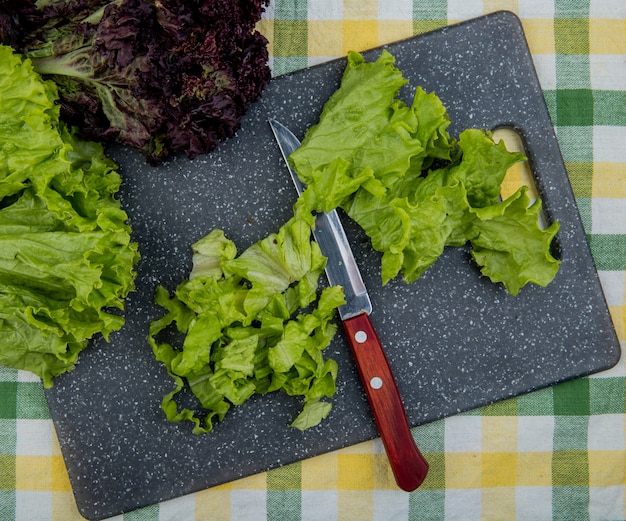 Vista superior de lechuga cortada con un cuchillo en la tabla de cortar y una entera con albahaca sobre tela escocesa