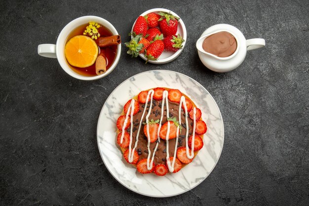 Vista superior desde lejos apetitoso pastel una taza de té con limón y canela junto a los tazones de fresas y crema de chocolate en el centro de la mesa