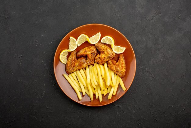 Vista superior desde lejos comida en alitas de pollo de placa con papas fritas y limón en un plato naranja sobre la mesa oscura