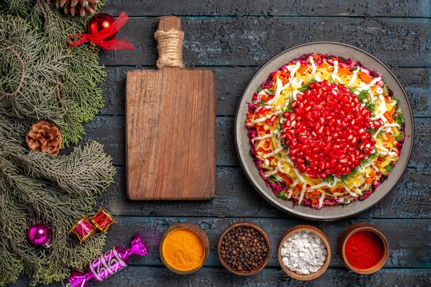 Vista superior desde lejos comida navideña Plato navideño con granada, tazones de fuente de tabla de cortar de especias y ramas de abeto con juguetes de árbol de Navidad