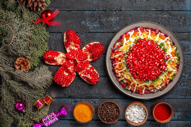 Vista superior desde lejos comida navideña Plato navideño con granadas tazones de especias, granadas y ramas de abeto apiladas con juguetes para árboles de Navidad