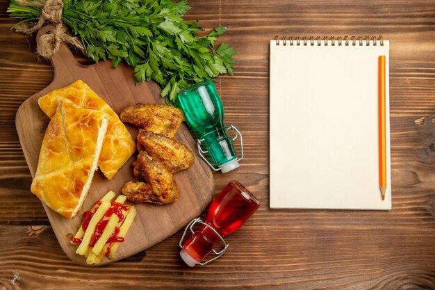 Vista superior desde lejos, comida rápida, hierbas, papas fritas, pollo y trozos de pastel en la tabla de cortar junto a las botellas de lápiz de cuaderno blanco y hierbas