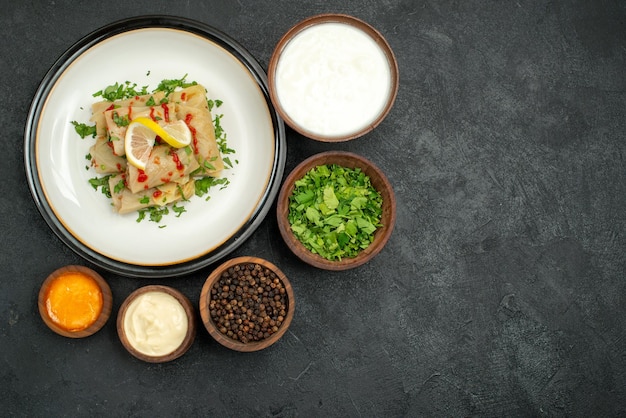 Vista superior desde lejos comida en tazones de mesa de crema agria hierbas pimienta negra y salsa amarilla y repollo relleno con hierbas limón y salsa en plato blanco sobre mesa negra