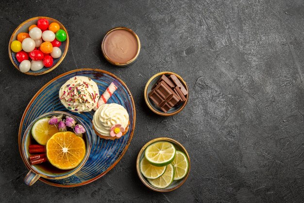 Vista superior desde lejos dulces sobre la mesa cuencos de caramelos chocolate crema de chocolate y rodajas de limas junto al plato de dos cupcakes y la taza de té de hierbas sobre la mesa