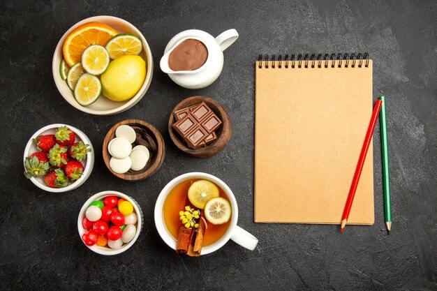 Vista superior desde lejos dulces sobre la mesa una taza de té con canela y limón junto al cuaderno de crema con lápiz verde y rojo y tazones de fresas, chocolate y caramelos