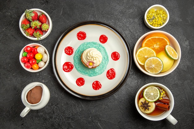 Foto gratuita vista superior desde lejos dulces con té una taza de té con anís estrellado junto al plato de cupcake y los cuencos de hierbas, frutas cítricas, chocolate, crema, fresas sobre la mesa