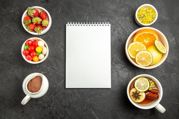 Vista superior desde lejos dulces con té una taza de té con canela y anís estrellado junto al cuaderno blanco y los tazones de hierbas, frutas cítricas, chocolate, crema, fresas sobre la mesa