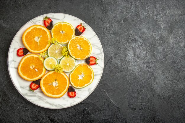 Vista superior desde lejos fresas cubiertas de chocolate naranja y limón en rodajas de naranja limón en un plato blanco sobre la mesa oscura