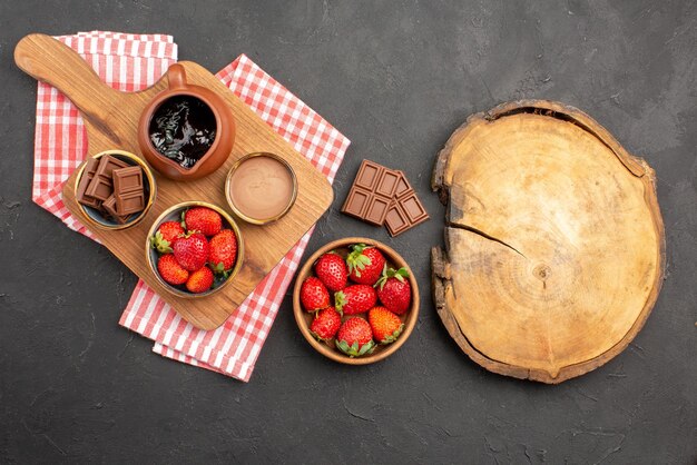 Vista superior desde lejos fresas y fresas con chocolate crema de chocolate en un tazón sobre la tabla de cortar sobre el mantel junto al plato de fresas y tabla de cortar