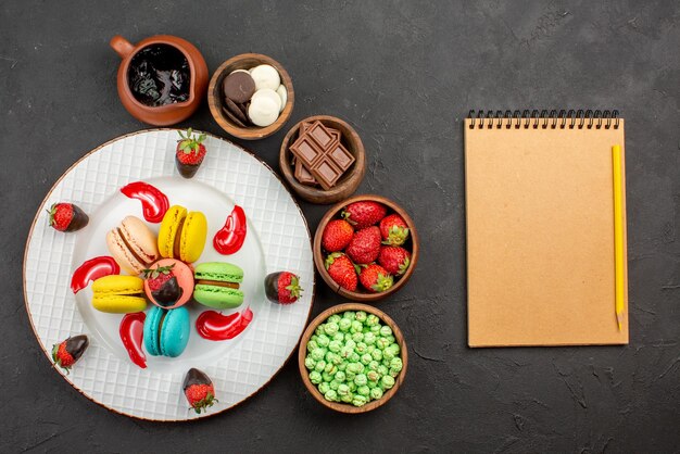 Vista superior desde lejos fresas y macarrones plato de fresas cubiertas de chocolate Macarrones franceses cuencos de dulces a su alrededor y cuaderno con lápiz sobre la mesa