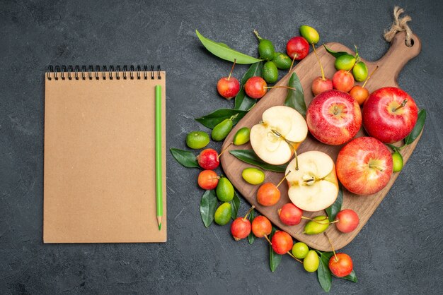 Vista superior desde lejos frutas manzanas cítricos en la tabla de cortar cerezas lápiz de cuaderno