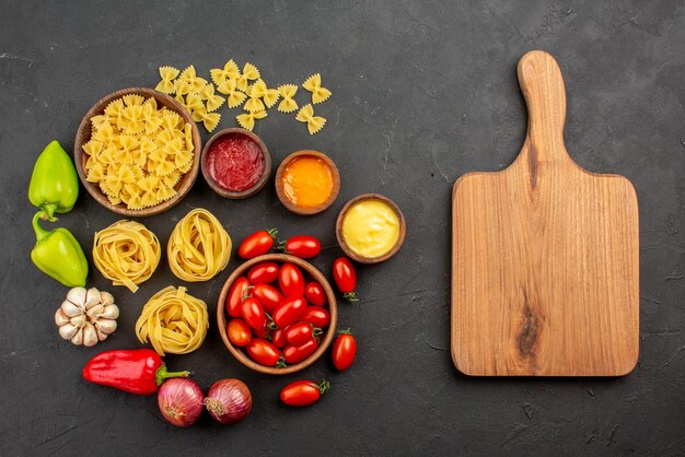 Vista superior desde lejos pasta y salsas tomates y pasta en tazones pimiento ajo tres tipos de salsas cebolla junto a la tabla de cortar sobre la mesa