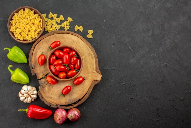 Vista superior desde lejos pasta y tomates cuenco marrón de tomates en la tabla de madera pasta y cebolla pimiento y ajo en el lado izquierdo de la mesa