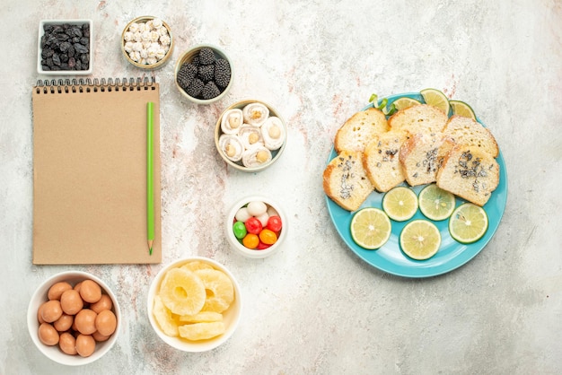 Vista superior desde lejos pastel de limón siete tazones de dulces junto al cuaderno de crema con lápiz verde plato de trozos de pastel con limón sobre la mesa