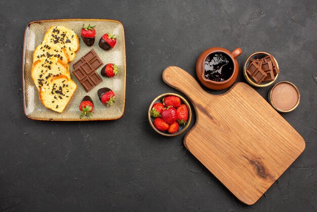 Vista superior desde lejos pastel de postre con fresas cubiertas de chocolate y chocolate y tabla de cortar entre tazones de crema de chocolate y bayas sobre la mesa