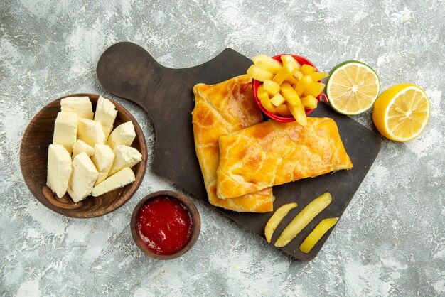 Vista superior desde lejos queso papas fritas tazón de queso salsa de tomate limón y papas fritas y apetitosos pasteles en el tablero de la cocina en la mesa gris