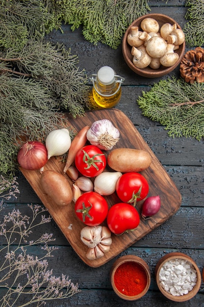 Vista superior desde lejos tabla de cortar de verduras y ramas con verduras entre especias coloridas y tazón de aceite de champiñones blancos y ramas de abeto con conos