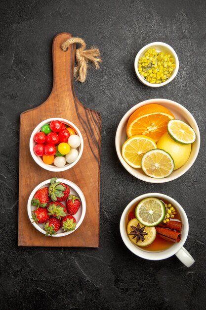 Vista superior desde lejos una taza de té, caramelos de colores y fresas en la tabla de cortar de madera junto a los cuencos de hierbas y limones en rodajas y una taza de té