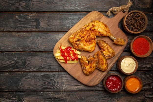 Vista superior desde lejos, tazones de muslo y alitas de pollo con salsas y especias coloridas y muslo y alas de pollo y papas fritas en la tabla de cortar