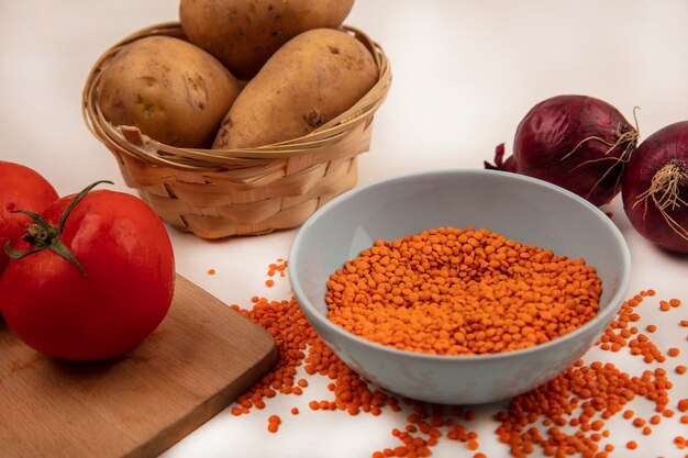 Vista superior de lentejas naranjas en un cuenco con patatas en un balde con tomates en una tabla de cocina de madera con cebollas rojas aisladas en una pared blanca