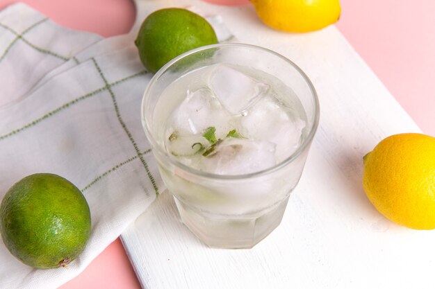 Vista superior de limonada fría fresca con hielo dentro del vaso junto con limones frescos en la superficie rosa