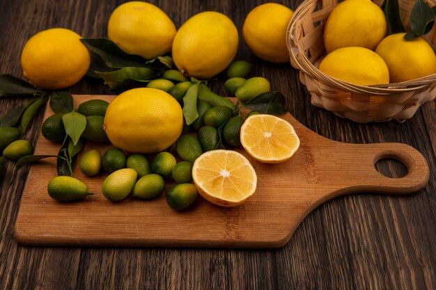 Vista superior de limones amarillos en un balde con limones y kinkans en una tabla de cocina de madera sobre una superficie de madera