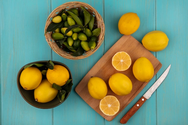 Vista superior de limones de forma redondeada en un recipiente con limones aislado en una tabla de cocina de madera con cuchillo con kinkans en un cubo en una pared de madera azul