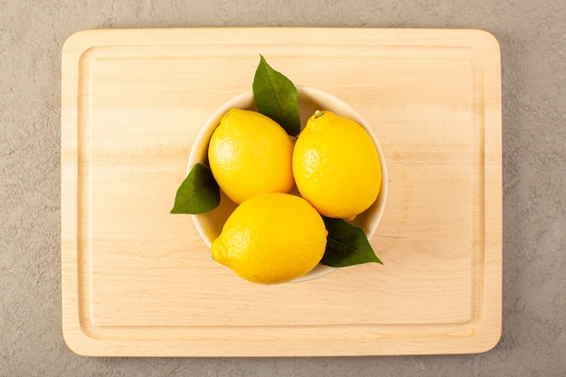 Una vista superior limones frescos amarillos maduros jugosos suaves con hojas verdes dentro de un tazón blanco forrado en el fondo gris frutas color cítrico