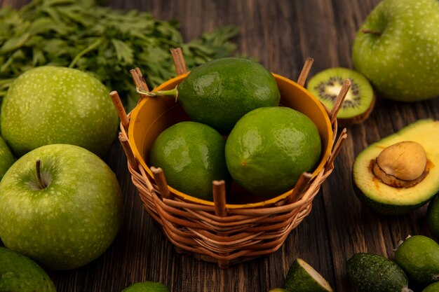 Vista superior de limones frescos en un balde con manzanas verdes kiwi feijoas aguacates y perejil aislado en una pared de madera