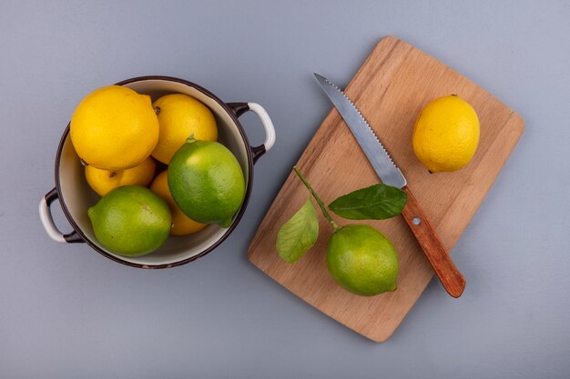 Vista superior de limones con limas en una cacerola con un cuchillo sobre una tabla de cortar sobre un fondo gris