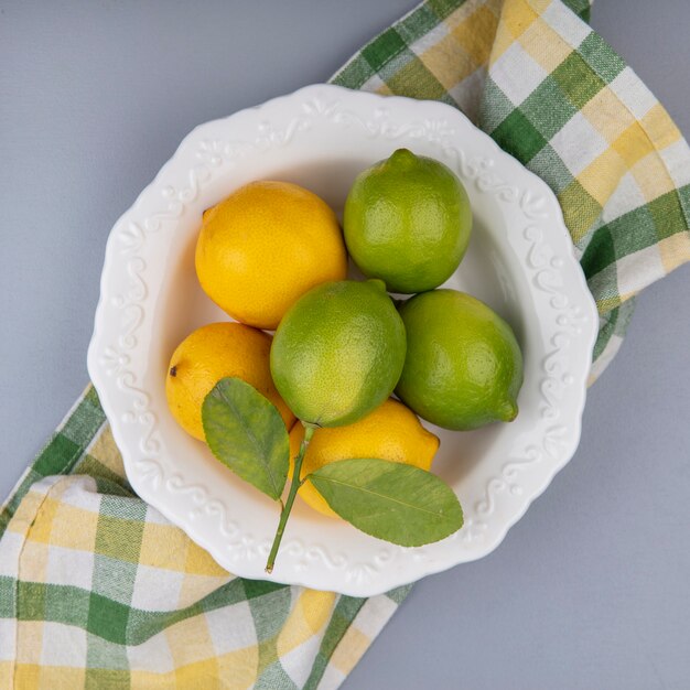 Vista superior de limones con limas en un plato sobre una toalla a cuadros amarilla sobre un fondo gris