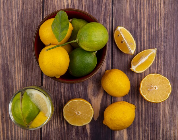 Vista superior de limones con limas en un tazón y un vaso de agua de desintoxicación sobre fondo de madera