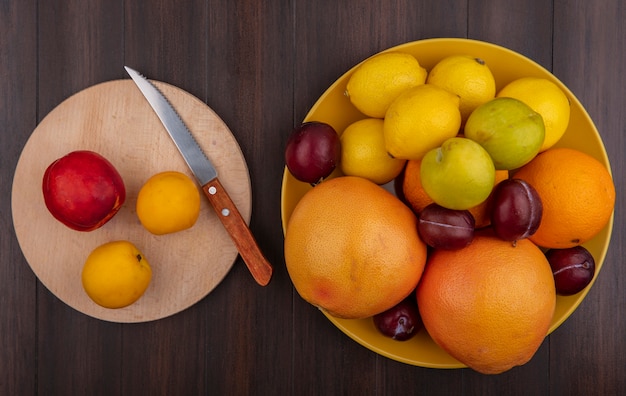 Vista superior de limones con naranjas, ciruelas y pomelo en un recipiente amarillo con albaricoques y melocotón en un soporte con un cuchillo sobre un fondo de madera