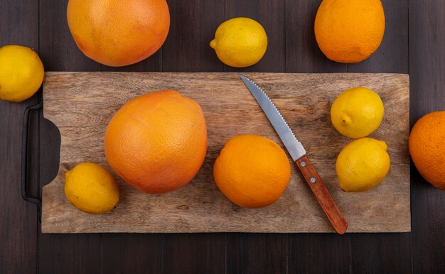 Vista superior de limones con naranjas y pomelos en una tabla de cortar con un cuchillo sobre un fondo de madera