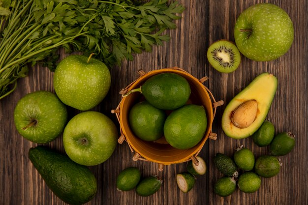 Vista superior de limones verdes en un balde con manzanas kiwi feijoas aguacates y perejil aislado en una pared de madera