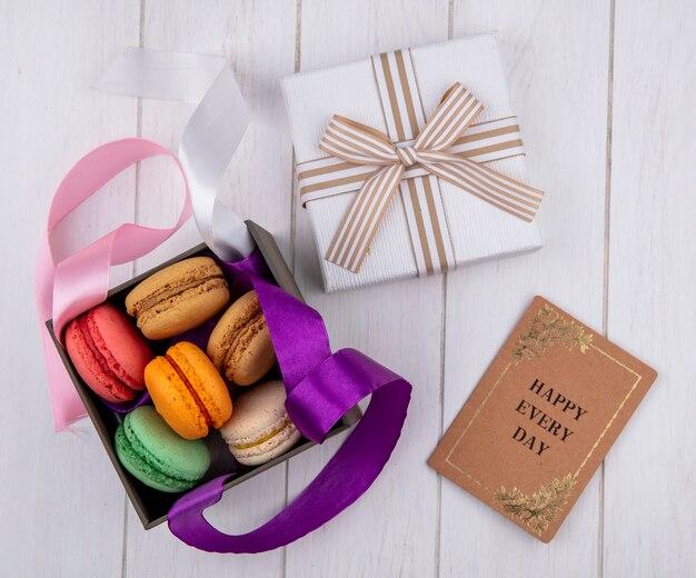 Vista superior de macarons de colores en una caja con lazos de colores y una caja de regalo con un libro sobre una superficie blanca