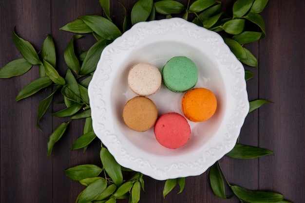 Foto gratuita vista superior de macarons de colores en un plato con ramas de hojas sobre una superficie de madera