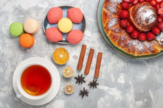 Vista superior de macarons franceses con pastelitos pastel de fresa y una taza de té en la superficie blanca pastel galleta azúcar pastel dulce té