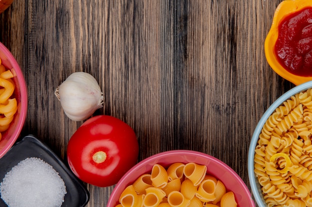 Foto gratuita vista superior de macarrones como rotini pipe-rigate y otros en tazones con ketchup, sal, ajo, tomate sobre madera