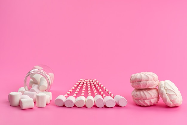 Una vista superior de malvaviscos blancos con palos rosas junto con merengues en el escritorio rosa, color dulce de azúcar