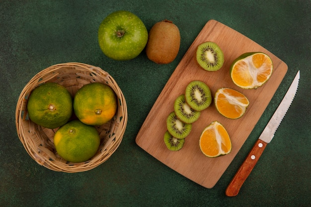 Vista superior de mandarinas en canasta con rodajas de kiwi en tabla de cortar con manzana