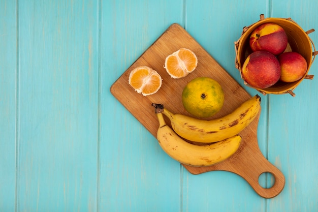 Vista superior de mandarinas enteras y medias en una tabla de cocina de madera con plátanos con melocotones en un balde en una pared de madera azul con espacio de copia