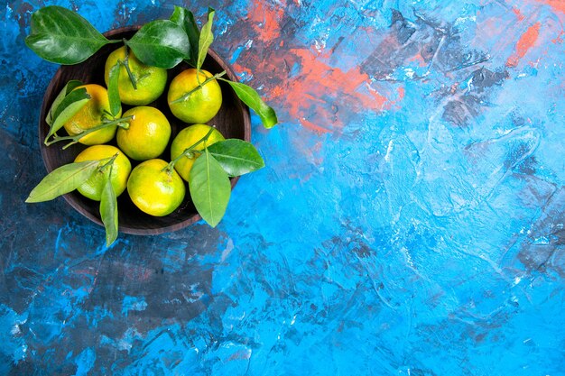 Vista superior de mandarinas frescas con hojas en un tazón de madera sobre una superficie azul aislada con espacio de copia