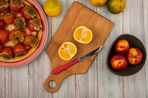 Vista superior de mandarinas frescas orgánicas en una tabla de cocina de madera con cuchillo con melocotones en un recipiente con caquis en un plato sobre un fondo de madera gris