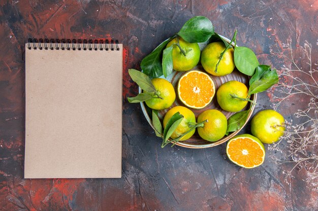 Vista superior de mandarinas frescas en un tazón un cuaderno sobre una superficie de color rojo oscuro